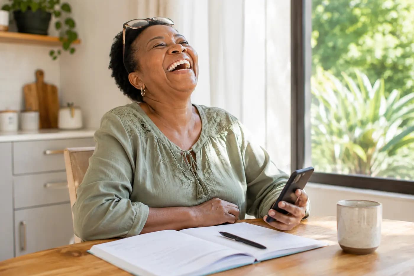 A grandmother laughing at the kitchen table, phone in hand, in bright daylight