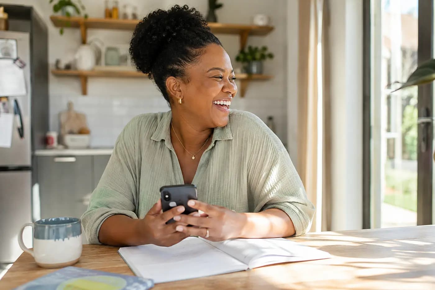 A mother laughing at the kitchen table, phone in hand, morning light