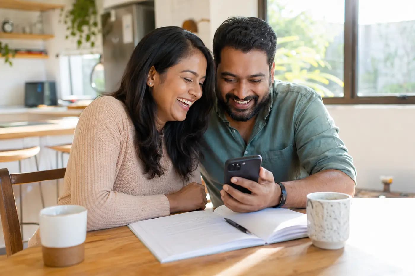Two parents laughing together over one phone in a sunlit kitchen