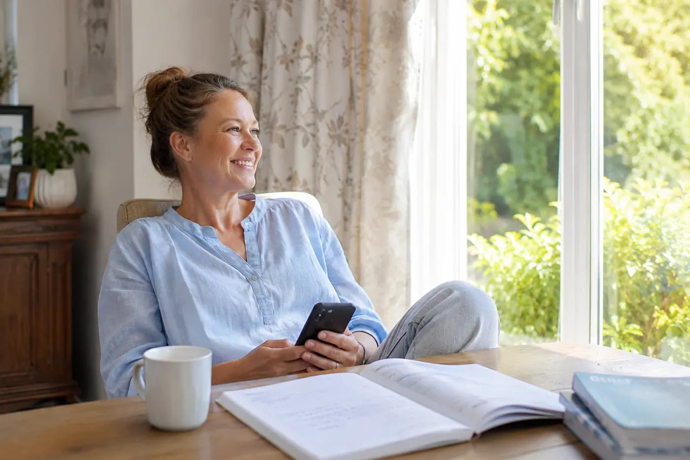 A mother smiling by a bright window, phone in hand, exercise book open